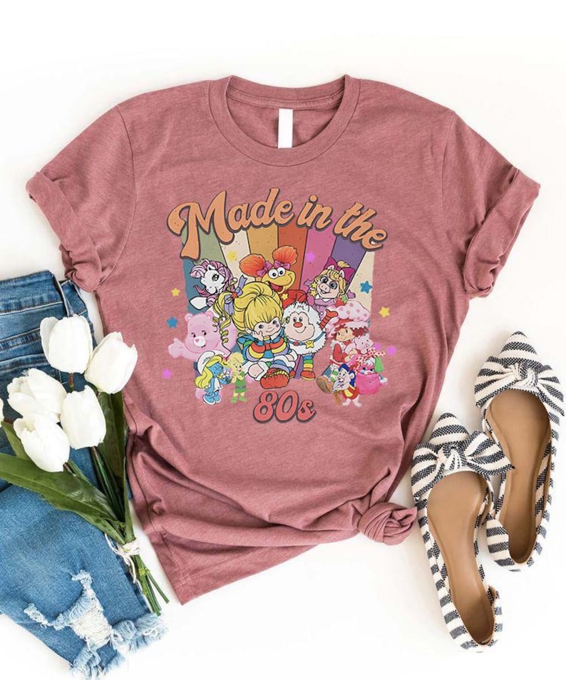May include: A dusty rose t-shirt featuring a colorful graphic of cartoon characters with the text "Made in the 80s". The design includes a rainbow background. A pair of striped flats and a bouquet of white flowers are also in the image.