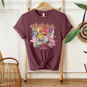 May include: Burgundy t-shirt featuring a colorful graphic with the text "Made in the 80s." The graphic includes cartoon characters and a rainbow design. The shirt is knotted at the waist and hangs on a wooden hanger.