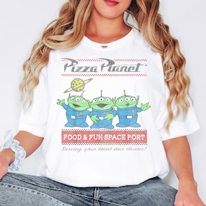 Puede incluir: Camiseta blanca con el logo de Pizza Planet y tres extraterrestres verdes con trajes espaciales azules. La camiseta tambi&eacute;n incluye el texto "FOOD & FUN SPACE PORT" y "Serving your local star cluster!"