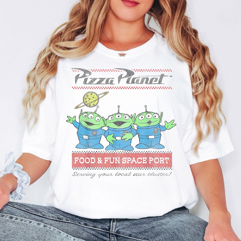Puede incluir: Camiseta blanca con el logo de Pizza Planet y tres extraterrestres verdes con trajes espaciales azules. La camiseta tambi&eacute;n incluye el texto "FOOD & FUN SPACE PORT" y "Serving your local star cluster!"
