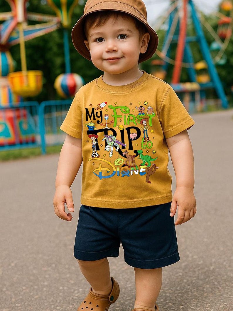 May include: A young child wearing a mustard yellow t-shirt and navy blue shorts. The t-shirt features the text "My First Trip to Disney" with cartoon characters. The child is also wearing a brown bucket hat.
