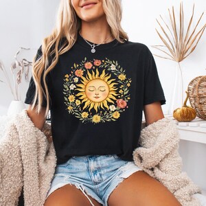 May include: Black t-shirt featuring a sun design with a smiling face, surrounded by a floral wreath. The sun is yellow with a light skin tone, and the flowers are in shades of pink, orange, and yellow. The shirt is a casual style.