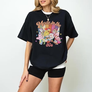 May include: Black t-shirt with the text "Made in the 80s" and a colorful graphic of cartoon characters. The shirt has short sleeves and a crew neck. The graphic includes rainbows, stars, and various cartoon figures.