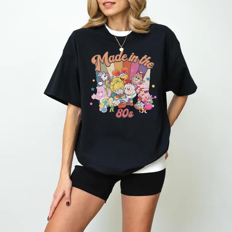 May include: Black t-shirt with the text "Made in the 80s" and a colorful graphic of cartoon characters. The shirt has short sleeves and a crew neck. The graphic includes rainbows, stars, and various cartoon figures.
