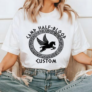 May include: White t-shirt featuring a black graphic of a winged horse inside a circular design with the words "CAMP HALF-BLOOD" and "CUSTOM". The design is in a Greek key style. The shirt is made of a soft material.