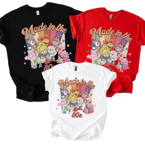 May include: Three t-shirts in black, red, and white, each featuring a retro design. The design includes the text "Made in the 80s" in a curved font, with a colorful rainbow background and cartoon characters. The shirts are short-sleeved.