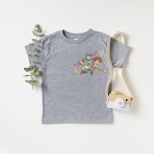 May include: A heather gray t-shirt featuring a colorful graphic of Buzz Lightyear and Woody from Toy Story. The cartoon characters are depicted in mid-air. A small, wooden toy camera with a white strap is also in the image.