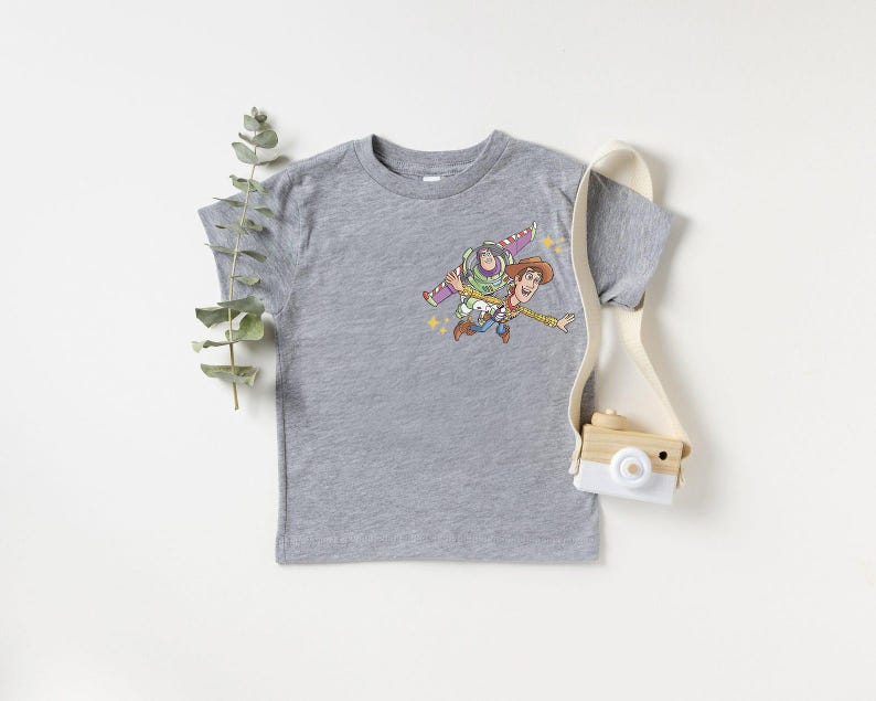 May include: A heather gray t-shirt featuring a colorful graphic of Buzz Lightyear and Woody from Toy Story. The cartoon characters are depicted in mid-air. A small, wooden toy camera with a white strap is also in the image.
