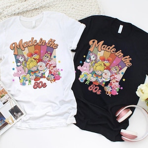May include: Two t-shirts, one white and one black, featuring a colorful graphic design. The design includes the text "Made in the 80s" with a rainbow background and cartoon characters. The shirts are short-sleeved and have a crew neck.