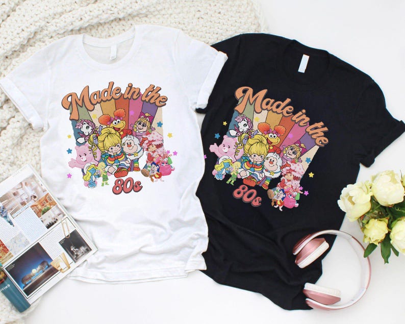 May include: Two t-shirts, one white and one black, featuring a colorful graphic design. The design includes the text "Made in the 80s" with a rainbow background and cartoon characters. The shirts are short-sleeved and have a crew neck.