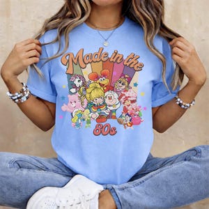 May include: Light blue t-shirt with a graphic that says "Made in the 80s." The graphic features colorful cartoon characters and a rainbow design. The shirt is a short-sleeved crew neck.