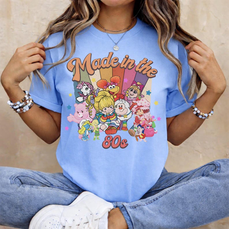 May include: Light blue t-shirt with a graphic that says "Made in the 80s." The graphic features colorful cartoon characters and a rainbow design. The shirt is a short-sleeved crew neck.