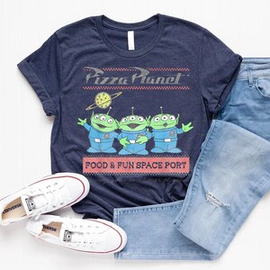 Puede incluir: Camiseta azul marino con el logo de Pizza Planet y tres personajes alien&iacute;genas verdes. La camiseta tambi&eacute;n tiene el texto "FOOD & FUN SPACE PORT". Zapatillas blancas y vaqueros azules tambi&eacute;n est&aacute;n en la imagen.