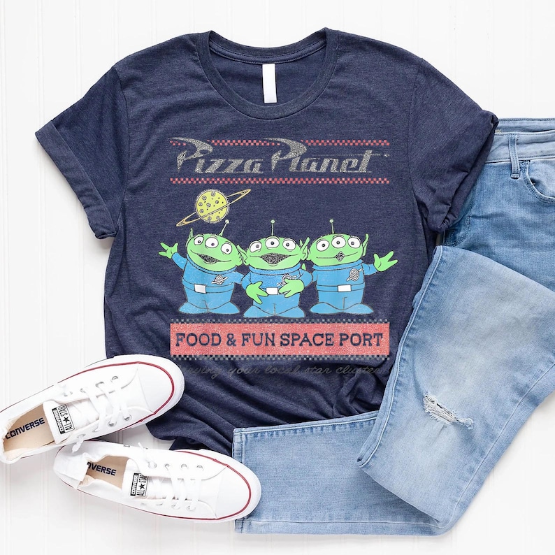 Puede incluir: Camiseta azul marino con el logo de Pizza Planet y tres personajes alien&iacute;genas verdes. La camiseta tambi&eacute;n tiene el texto "FOOD & FUN SPACE PORT". Zapatillas blancas y vaqueros azules tambi&eacute;n est&aacute;n en la imagen.
