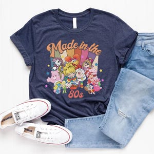 May include: Navy blue t-shirt with a colorful graphic featuring cartoon characters and the text "Made in the 80s." The shirt is paired with light blue jeans and white sneakers.