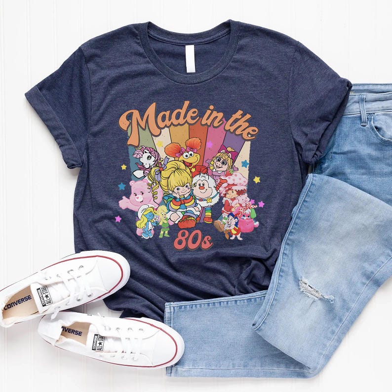 May include: Navy blue t-shirt with a colorful graphic featuring cartoon characters and the text "Made in the 80s." The shirt is paired with light blue jeans and white sneakers.