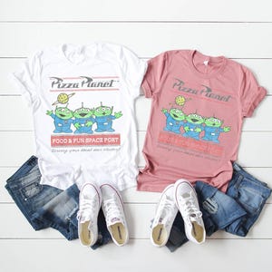 Puede incluir: Dos camisetas, una blanca y otra rosa polvoriento, con el logo de Pizza Planet y tres personajes alien&iacute;genas verdes. Las camisetas se combinan con vaqueros azules y zapatillas blancas Converse. El texto de las camisetas dice "FOOD & FUN SPACE PORT".