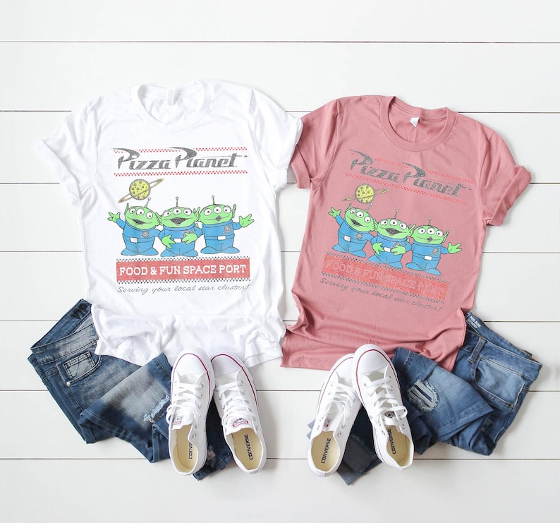 Puede incluir: Dos camisetas, una blanca y otra rosa polvoriento, con el logo de Pizza Planet y tres personajes alien&iacute;genas verdes. Las camisetas se combinan con vaqueros azules y zapatillas blancas Converse. El texto de las camisetas dice "FOOD & FUN SPACE PORT".