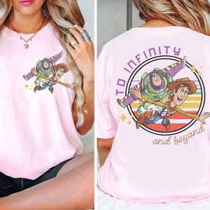 May include: A light pink t-shirt featuring a graphic of Buzz Lightyear and Woody from Toy Story. The front of the shirt shows the characters flying. The back of the shirt has the phrase "To infinity and beyond" in a circle.