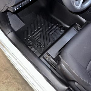 May include: Black car floor mats with a textured, geometric design. The mats are fitted in a vehicle, visible near the driver's seat and dashboard. The mats appear to be made of a durable, weather-resistant material.