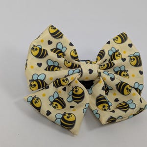 Peut inclure: Un nœud pour les cheveux crème avec un motif répété d'abeilles de dessin animé et de petits cœurs noirs. Les abeilles sont jaunes et noires avec des ailes bleues. Le nœud est noué en forme de nœud classique.