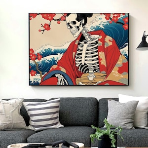 以下が含まれることがあります： 赤い着物を着た骸骨が、青い波と桜を背景にした額装されたアートワーク。アート作品は黒いフレームで、灰色のソファの上に飾られています。