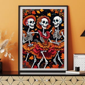 以下が含まれることがあります： カラフルな衣装を着た3体の踊る骸骨が描かれた額入りのアートプリント。骸骨はオレンジと赤の色合いの帽子とフリルスカートを身に着け、秋の葉が描かれた黒い背景に配置されています。このアートワークは、死者の日のテーマです。