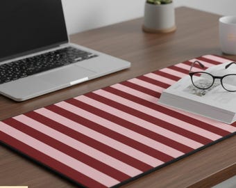 May include: A rectangular desk mat with a striped pattern in alternating shades of red and pink. A laptop, a book, and glasses rest on the mat, which is placed on a wooden desk. A small succulent plant in a gray pot is also visible.