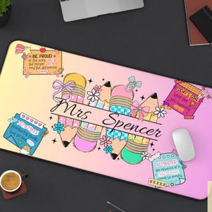 May include: A personalized desk mat with a gradient yellow to pink background. The mat features pencil illustrations, floral accents, and the name "Mrs Spencer" in a script font. Inspirational quotes are also included.