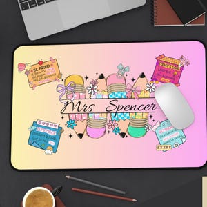 May include: A personalized mousepad with a gradient pink and yellow background. The design features colorful pencils, flowers, and the name "Mrs. Spencer" in script. Small notes with text are scattered around the design.