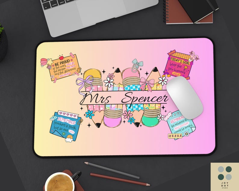 May include: A personalized mousepad with a gradient pink and yellow background. The design features colorful pencils, flowers, and the name "Mrs. Spencer" in script. Small notes with text are scattered around the design.