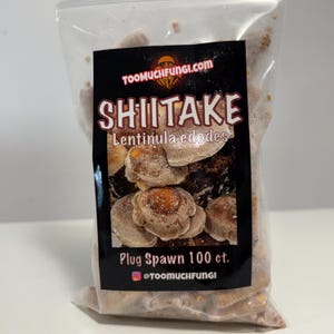 May include: A clear plastic bag filled with shiitake mushrooms. A black label displays the text "TOOMUCHFUNGI.COM" and "SHIITAKE Lentunila edodes". Below, the label reads "Plug Spawn 100 ct." and "@TOOMUCHFUNGI". The mushrooms are brown and beige.