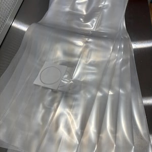 May include: A stack of clear, transparent plastic bags, possibly for packaging or storage. The bags are folded and stacked neatly on a reflective, metallic surface. A few white circular stickers are visible.