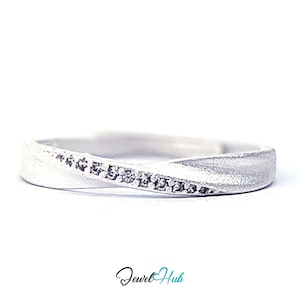 Op de afbeelding: Een zilverkleurige ring met een getextureerde, matte afwerking en een gedraaid bandontwerp. Een diagonaal kanaal bezet met kleine, heldere, sprankelende steentjes loopt over het midden van de band. De tekst Jewel Hub staat in zwart en blauwgroen eronder.