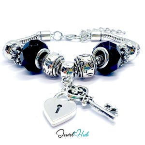 May include: A silver-tone snake chain charm bracelet featuring two faceted black beads, two silver beads with engraved patterns, and a central dangling charm set consisting of a heart-shaped padlock and a decorative skeleton key.