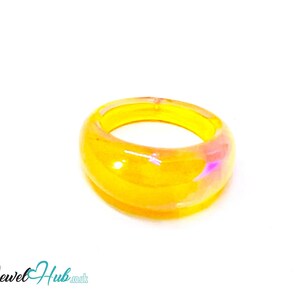May include: A chunky, translucent ring with a vibrant yellow hue. The ring has a rounded shape and a smooth, glossy finish. Hints of pink and purple shimmer within the material, adding depth and visual interest. The ring is set against a clean white background.