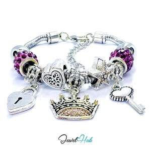 May include: A silver-tone charm bracelet featuring two purple rhinestone-encrusted beads, a heart-shaped padlock charm, a crown charm, a star charm, and a heart-topped key charm. The bracelet includes a metal chain with a lobster claw clasp.