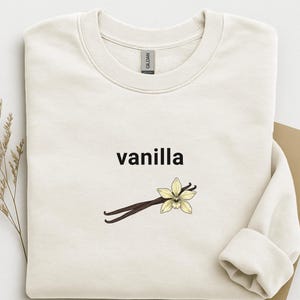 Puede incluir: Sudadera color crema con la palabra "vanilla" en negro, sobre un gráfico de una flor de vainilla y dos vainas de vainilla marrones. La sudadera tiene cuello redondo y mangas largas.