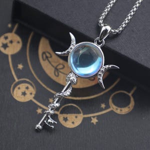 May include: A silver pendant necklace with a celestial design. The pendant features an iridescent orb, crescent moons, and a staff-like element with a snake motif. The necklace is displayed on a dark surface.