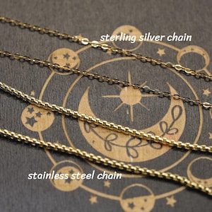 May include: A silver-colored chain labeled sterling silver chain and a gold-colored chain labeled stainless steel chain. The chains are displayed against a dark background featuring a circular celestial design with a crescent moon and stars.
