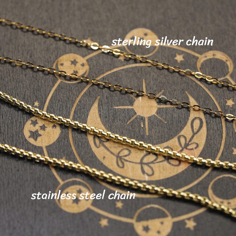 May include: A silver-colored chain labeled sterling silver chain and a gold-colored chain labeled stainless steel chain. The chains are displayed against a dark background featuring a circular celestial design with a crescent moon and stars.