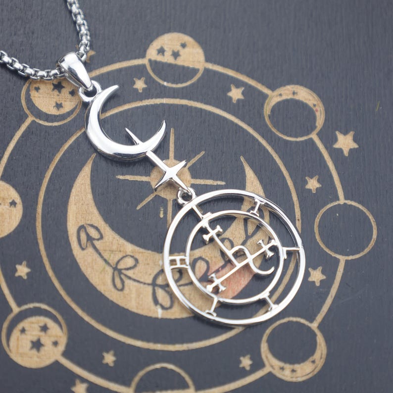 May include: Silver-toned pendant necklace featuring a crescent moon, star, and a circular emblem with intricate symbols. The necklace hangs on a silver chain. The background is dark with celestial-themed illustrations.