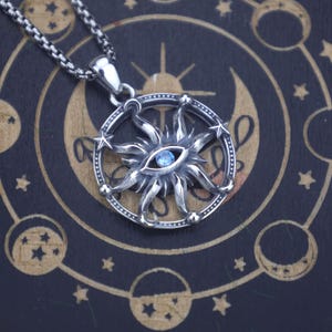 Puede incluir: Un collar con colgante plateado con un diseño de sol y un ojo central de piedra preciosa azul. El colgante está detallado con estrellas y un marco circular, suspendido de una cadena plateada. El fondo tiene un patrón celestial.