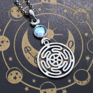 May include: A silver-toned pendant necklace featuring a circular labyrinth design with a central six-pointed star motif. Above the labyrinth, a small crescent moon connects to a round, iridescent blue cabochon stone, topped by another crescent.