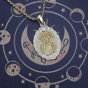 May include: A gold-toned oval pendant necklace featuring a central embossed figure of a deity with multiple arms, surrounded by a border of small clear stones and a single round iridescent stone at the top, suspended from a gold-toned box chain.