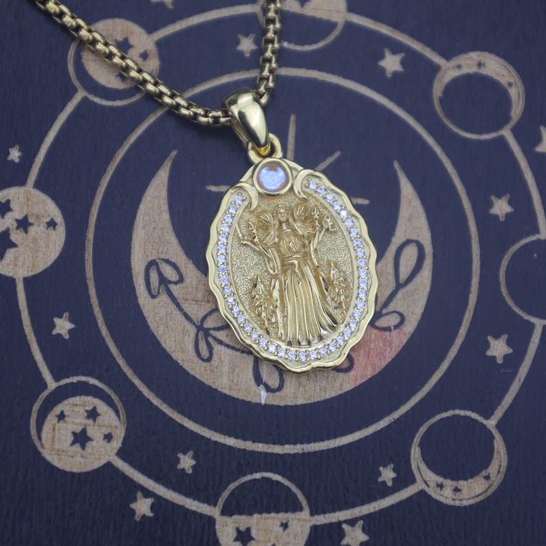 May include: A gold-toned oval pendant necklace featuring a central embossed figure of a deity with multiple arms, surrounded by a border of small clear stones and a single round iridescent stone at the top, suspended from a gold-toned box chain.