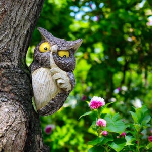 May include: A whimsical owl sculpture, partially embedded in a tree trunk, with a finger pressed to its beak in a 'shush' gesture. The owl has yellow eyes and a textured, brown and gray finish. The background features lush green foliage and pink flowers.