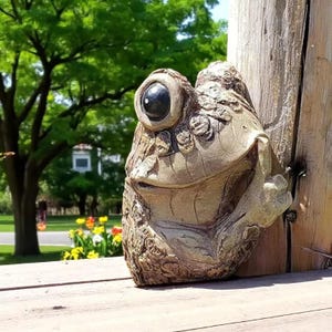 May include: A detailed, textured frog sculpture with a large, dark eye and a smiling expression. The frog is attached to a wooden post, with a natural, earthy color palette. The sculpture is likely for outdoor garden decor.