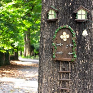 Puede incluir: Una caprichosa puerta de hadas y dos decoraciones de ventana están unidas a un tronco de árbol. La puerta marrón tiene un diseño de flor y herrajes plateados. Una pequeña escalera de madera cuelga debajo.