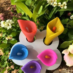 Puede incluir: Un conjunto de velas de cala de colores vibrantes: rojo, amarillo, azul, rosa y morado. Las velas están dispuestas en un soporte blanco con forma de flor, rodeadas de vegetación y flores blancas. Un artículo decorativo.